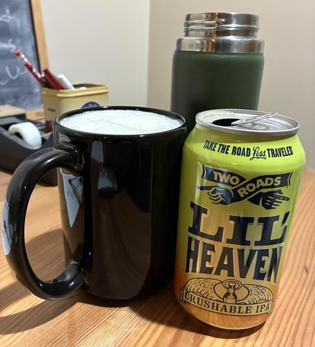 A beer in a black coffee cup with the beer can on the side. There is an open insulated coffee mug in the back.