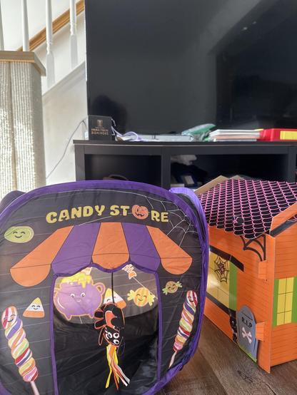 Children's Halloween-themed play tents on a wooden floor. The tent in the foreground has a black, purple, and orange color scheme with "CANDY STORE" written on it. There are illustrations of candy, a purple teapot, smiling candies, and a hanging plush spider in the entrance. To the right, there is another tent resembling a haunted house, decorated in orange with a roof pattern, spider webs, and a gravestone marked "RIP." In the background, there's a television on an entertainment stand, some books or papers, and a staircase with white spindles and a wooden banister.

Image description provided with Be My Eyes.