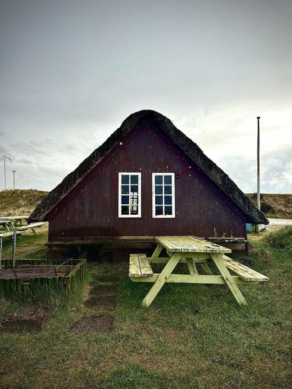 A traditional small house with a steep, thatched roof and dark wood siding stands raised on a foundation above a grassy area. In front of the house is a weathered wooden picnic table with attached benches. A gravel path leads up to the house under an overcast sky.