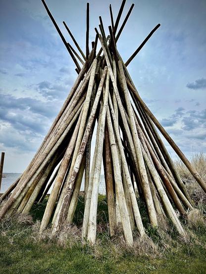 Large teepee-like structure made of numerous long, weathered wooden poles, arranged pointedly upwards. The structure is set against a cloudy sky with a glimpse of water visible in the background, surrounded by grassy terrain.