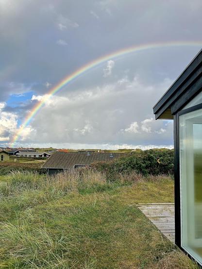 Vibrant rainbow arcs across a cloudy sky over a rural landscape. The right foreground features the corner of a modern building with large windows. Below the rainbow, a cluster of small, dark-roofed houses is nestled among grassy dunes. A wooden walkway leads towards one of the houses, surrounded by overgrown grass and wild foliage.