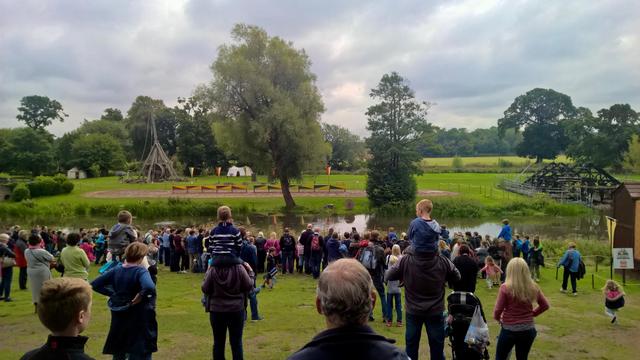 Warwick Castle in Warwick, England, overlooking a small pond beside an open field. Visitors, including families with children on shoulders and in strollers, watch a demonstration area featuring a large medieval trebuchet on the left, with colorful banners and a few white tents in the background. To the right, a wooden structure spans the water, and the landscape beyond is filled with green fields and clusters of trees under an overcast sky. The scene reflects a public outdoor event at the historic site, combining medieval displays with natural surroundings.