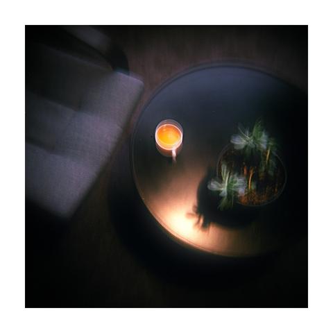 A high-angle, low-light photo with intentional motion blur shows a small potted plant and a glowing candle on a round, dark table. A rectangle of soft light falls on the chair to the left.