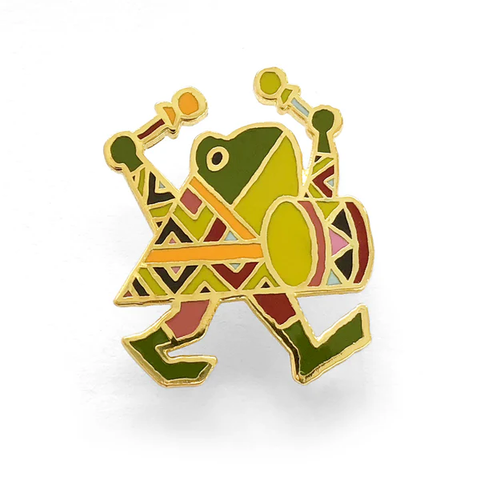 Photo, on a white background, of an enamel pin of an anthropomorphic frog, dressed in brown leggings peeking out from a triangular top, near enough wide at the hem as the frog is tall, decorated with geometric pattern reminiscent of Aztec design.

The figure appears to be mid-stride, walking to the right, face in profile, with both arms upraised, each flourishing a percussion mallet with heads of tied-off fabric, poised as if to strike either side of a marching bass drum, in front of them, in a position suggestive of a croaking frog's inflated vocal sac, the drum continuing the same pattern geometric pattern as their dress.
