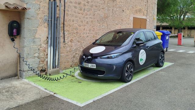 Un cotxe elèctric compartit de la cooperativa Ecotxe carrega electricitat d'un punt de càrrega a una paret d'un edifici antic. La plaça d'aparcament està pintada en verd.