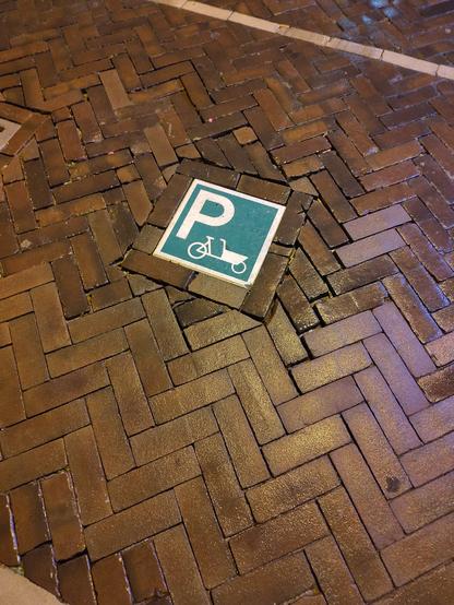 Herringbone pattern brick pavement with a green and white parking sign inlaid. As well as a large P there is a simplified cargo bike on the sign