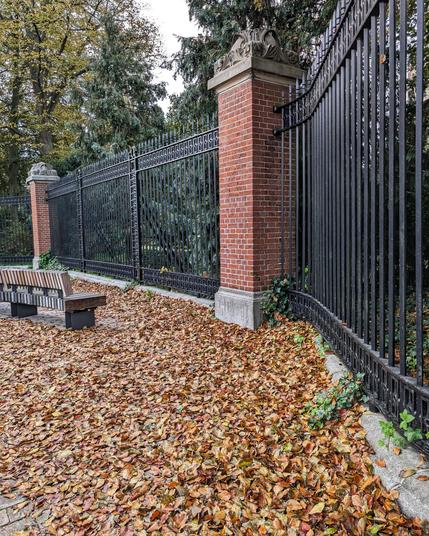 Pavement covered with crisp brown leaves. On the right hand side a large iron fence with brick posts, extended all along the right to left side of the photo.
In the middle of the left hand side is a wooden curving bench.