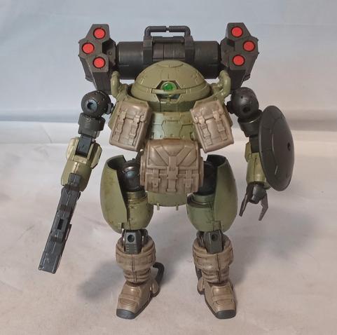 A green, black and beige mecha with a monoeye, three fingered claws, and rounded proportions. It has a gun, shield, backpack missiles and pouches on its chest. The pouches and lower legs seem to be fabric. Neutral pose.