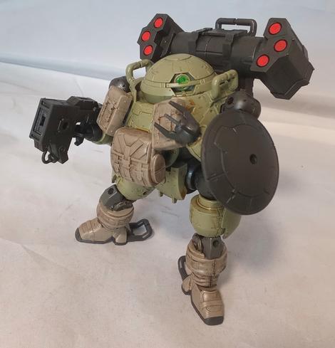 A green, black and beige mecha with a monoeye, three fingered claws, and rounded proportions. It has a gun, shield, backpack missiles and pouches on its chest. The pouches and lower legs seem to be fabric. Angled pose.
