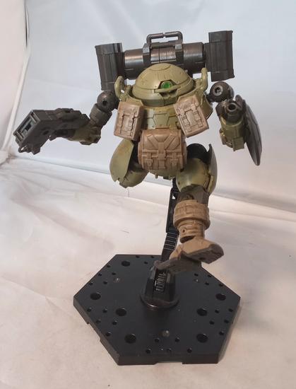 A green, black and beige mecha with a monoeye, three fingered claws, and rounded proportions. It has a gun, shield, backpack missiles and pouches on its chest. The pouches and lower legs seem to be fabric.  Leaping pose.
