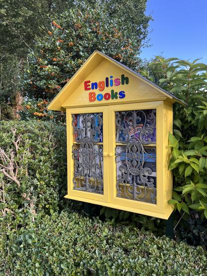 Yellow little library hidden in a hedge with iron decorative covers over the windows and English Books written at the top, where each letter has a different colour.