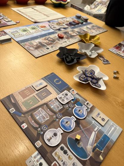 Ein Tisch mit verschiedene Spielmaterialien. Im Vordergrund ein Board für den Spieler, in der Mitte Schalen mit Klötzchen und im Hintergrund ein größeres Bord für das Spiel selbst. 

Das Thema ist wissenschaftlich.