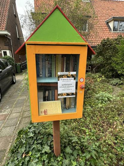 A colorful, small wooden bookcase with a green and red roof is displayed outdoors. It contains various books and features a sign promoting a workshop on communication. Surrounding the bookcase is greenery and a brick pathway, with a residential building in the background.