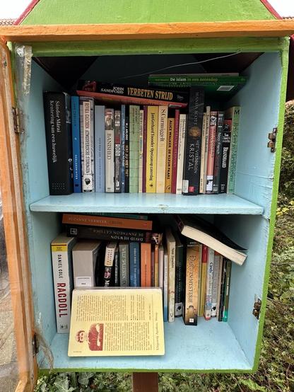 A small, colorful bookshelf is displayed outdoors, containing various titles across two shelves. The books cover a range of genres, including fiction and non-fiction, with some titles visible in Dutch.