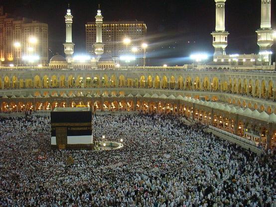 The Kaaba in Mecca, Saudi Arabia; nighttime photo of a large black cube surrounded by pilgrims within an open-air mosque; beyond are minarets & city buildings