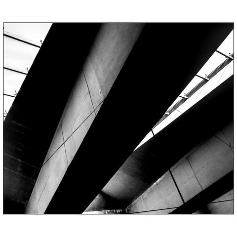This black-and-white photograph captures an abstract architectural scene, focusing on the geometric lines and angles of a modern structure. The image features a series of large, diagonal concrete beams and panels, creating a dynamic interplay of light and shadow. The light filters through narrow skylights or windows, casting sharp, linear shadows across the concrete surfaces. (draft by mistral.ai)
