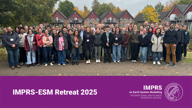 A group of about 70 scientists with badges around their necks gathered in a park-like area with similar holiday houses in the background. The leaves of the trees in the background are colored green, brown and yellow, which together with the jackets worn by most people suggests a chilly fall day.