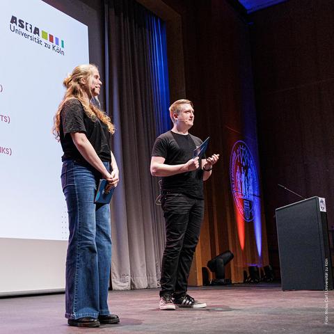 Zwei Personen stehen auf einer Bühne vor einer Präsentation mit dem Logo der Universität zu Köln.
