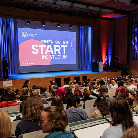 Ein gefüllter Hörsaal, auf der Bühne ist ein Bildschirm mit der Aufschrift 'Einen guten Start ins Studium!' sichtbar.