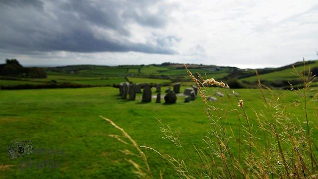 Stone circle in the middle of a grass field surrounded by rolling hills and dramatic clouds. Tall, dry grass in the foreground adds depth to the landscape scene.