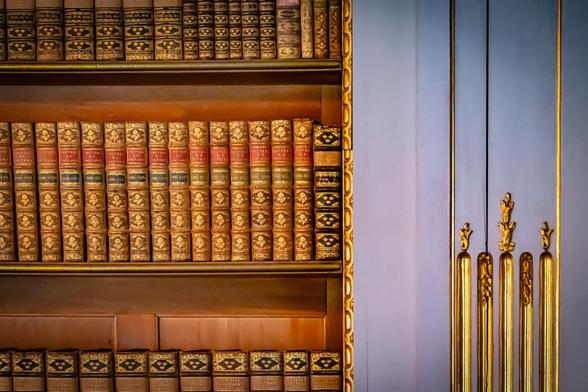 Ornate bookshelf filled with antique leather-bound books featuring gold and red detailing on spines. The bookshelf has intricate golden trim, with a section showing elaborate golden door handles on a purple background.