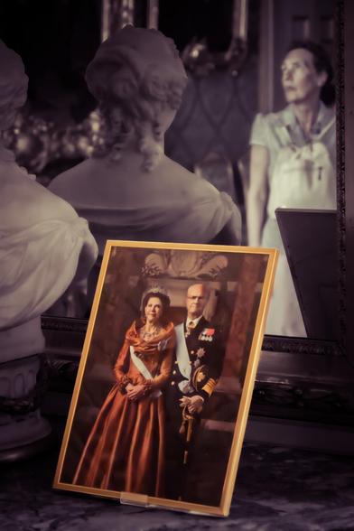 Framed portrait of a regal couple wearing formal attire with medals rests on a polished surface. A marble bust with detailed hair and clothing is positioned nearby, facing a mirror. The mirror reflects a woman in a uniform, partially blurred and standing in the background.