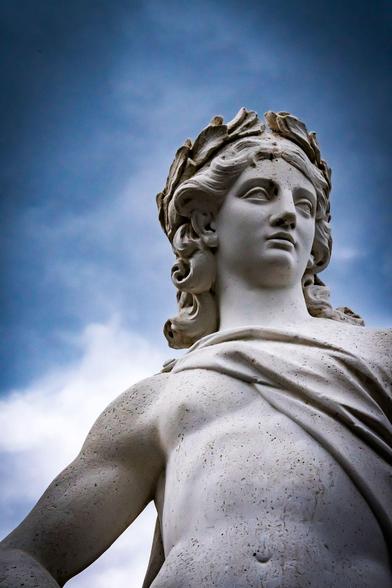 Marble statue of a classical male figure with a laurel wreath on his head, looking to the side. The statue is set against a backdrop of a cloudy blue sky, highlighting its detailed features and smooth texture. The figure's drapery is carved to fall across the shoulder, adding to the lifelike appearance.