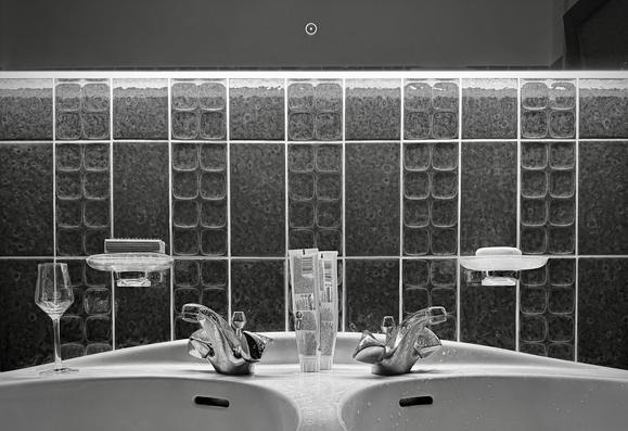das bild zeigt ein badezimmer-doppelwaschbecken mit zwei wasserhähnen.
zwischen den wasserhähnen stehen zwei tuben zahnpasta. rechts und links über dem waschbecken befinden sich seifenablagen mit einer fingerbürste und einem stück seife. links am rand des waschbeckens steht ein leeres spirituosenglas. die wand hinter dem waschbecken ist mit dunklen, strukturierten fliesen bedeckt, die in rechteckigen mustern angeordnet sind.
am oberen bildrand ist ein spiegel zu sehen. es hat mittig einen ein-aus-schalter auf der spiegelfläche und beleuchtet mit einem lichtstreifen stimmungsvoll die szene. das bild ist in schwarz-weiß aufgenommen.