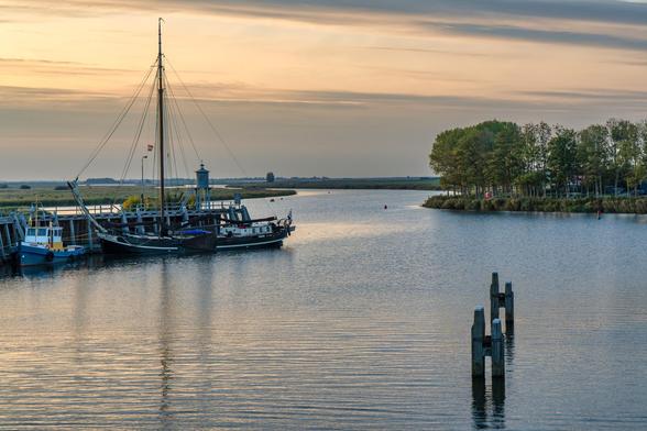 Die Szene zeigt eine weite Wasserlandschaft, die typischerweise auf die Umgebung von Zoutkamp am Lauwersmeer oder am Reitdiep hindeutet, aufgenommen in gedämpftem Abendlicht.
Vordergrund und Wasser:
Das Wasser ist ruhig und reflektiert den Himmel, wodurch es eine leicht silbrige bis bläuliche Farbe annimmt.
Im rechten Vordergrund ragen zwei dunkle, grobe Holzpfähle aus dem Wasser, wahrscheinlich Überreste einer alten Anlegestelle oder als Markierung dienend.
Boote und Anlegestelle (links):
Auf der linken Seite befindet sich eine Anlegestelle, an der mehrere Boote vertäut sind.
Das auffälligste Schiff ist ein traditionelles, dunkles Segelschiff mit einem hohen Mast, möglicherweise ein Tjalk oder ein Klipper der "Braunen Flotte" (traditionelle Segelboote für Charterfahrten).
Direkt davor liegt ein kleineres, weiß-blaues Beiboot oder Arbeitsboot.
Am Kai sind dunkle Holzbauten und eine Art Schleusen- oder Brückenwärterhaus mit einem Turm zu sehen.
Hintergrund und Landschaft:
Der Blick schweift über das offene Wasser, das sich in die Ferne erstreckt.
Am rechten Ufer befindet sich eine kleine Landzunge, bewachsen mit einer Gruppe von Bäumen, die ein frisches Grün aufweisen.
Am Horizont ist eine flache, weite Landschaft zu erkennen, die charakteristisch für die niederländische Region Groningen/Friesland ist, mit niedrigem, schilfartigem Bewuchs und vereinzelten, weit entfernten Silhouetten von Bäumen oder Gebäuden.
Eine rote Boje ist mittig im Wasser sichtbar.
Himmel und Licht:
Der 