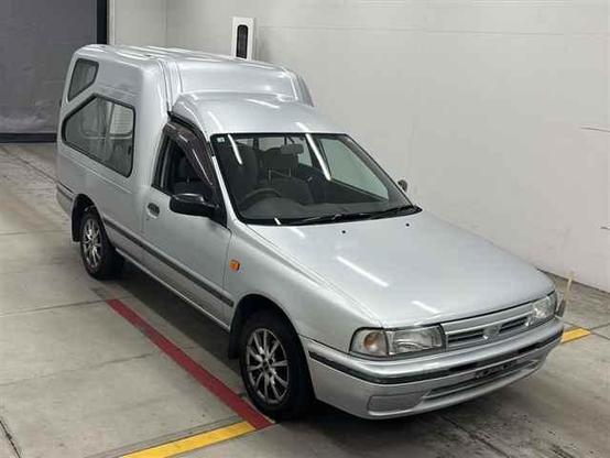 A silver Nissan AD Wagon MAX. A ute with a high-cube back instead of a bed. It has unusual geometric windows and dual french doors to get into the back.