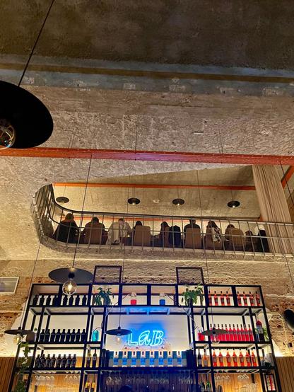 Dimly lit, multi-level bar with exposed concrete and brick. The view looks up at a curved balcony railing where people are silhouetted, with a well-stocked bar and a blue neon sign in the foreground.