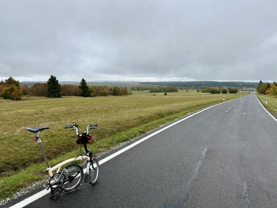 Brompton halb geklappt am Straßenrand. Regenwolken. Herbstfarben. Hochebene mit Rhön-typischen freien Flächen mit einzelnen Bäumen und Büschen. Im Hintergrund lässt sich das Schwarze Moor erahnen.
