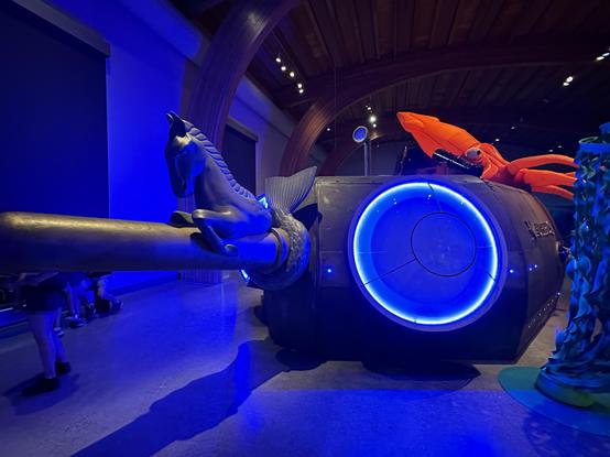 Color photo of a dimly blue-lit exhibit hall of a ‘20,000 Leagues Under the Sea’ inspired exhibit at Mystic Seaport, Mystic CT. A walk-through model of Captain Nemo’s Nautilus features a horse with the hind quarters of a fish — mermaid style — riding a long cylindrical prow jutting from the hull. The hull of the Nautilus is matte black, adult-height and about twice as wide, like a horizontally stretched cylinder. To the right of the prow is a large circular window — almost the height of the hull — circled by a bright blue ring of light. A brightly lit fluorescent orange giant squid rides on top of and grips the back of the hull.