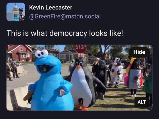 A group of people in inflatable costumes, including a blue Cookie Monster, a penguin, and a cow, are marching in a public area. Signs and banners are visible, and there are police officers in the background. The scene suggests a playful demonstration