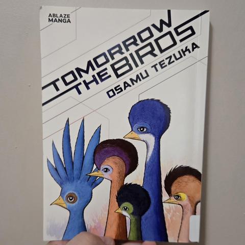 Photograph of Tomorrow the Birds against a lightly colored wall held by a white person's hand that is only partially visible at the bottom. The title of the series and the author (Osamu Tezuka) are both written in stylized text at an angle just above images of five busts / heads of various birds with enlarged tops of their heads to indicate larger brains. In the upper left hand, in small black print it reads "Ablaze Manga."