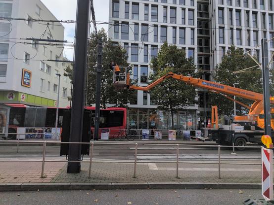 Eine Straßenbahnhaltestelle an der ein orangener Kran steht um die Oberleitung zu reparieren. Im Hintergrund ein roter Bus, und dahinter hohe, weißfarbene Häuser.