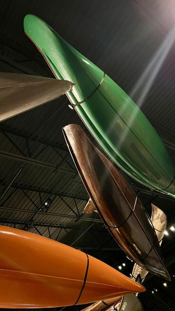 Canoes hang from the ceiling of a museum exhibit and it feels like you are below the swirling waters. Canoes are at various angles, two wooden, one orange and one green. A shaft of light shines diagonally across, and the ceiling is black.