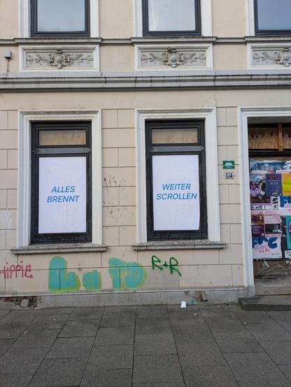 Ein leerstehendes Haus mitten in der Stadt, Türen und Fenster zugenagelt, getaggt und plakatiert. Auf die zwei Fensterscheiben hat jemand vollflächig und sehr akkurat weißes Papier geklebt. Das linke Fenster ist bedruckt mit den Worten „Alles brennt”, das rechte mkt „Weiter scrollen”.