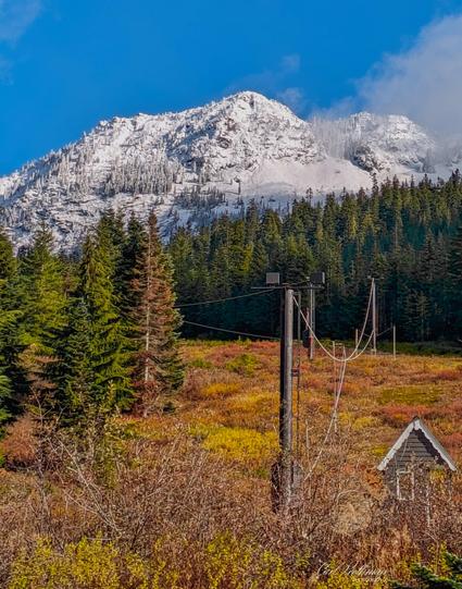A crisp fall morning at Snoqualmie Pass, Washington, featuring the historic Washington Alpine Club rope tow area. A rustic shack and weathered utility poles rest among colorful alpine meadow brush, framed by deep evergreen forest. Beyond, the first snow of the season dusts a rugged mountain ridge under a brilliant blue sky, evoking the quiet anticipation of winter’s return.
