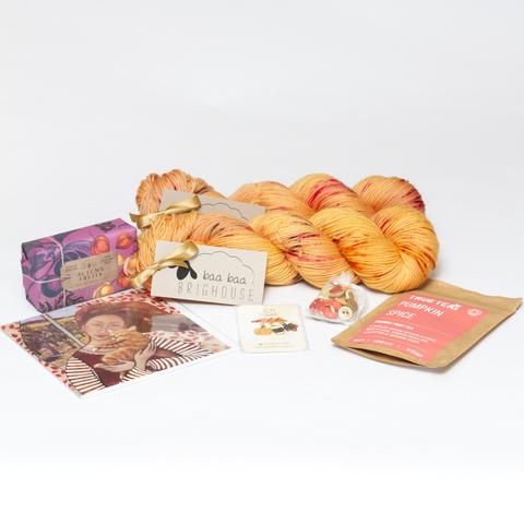 Picture shows two skeins of yarn, one behind the other, in shades of honey with red, purple and raspberry speckles. In front of the yarn, from left to right, is a soap, card, stitch markers, buttons and a packet of tea.