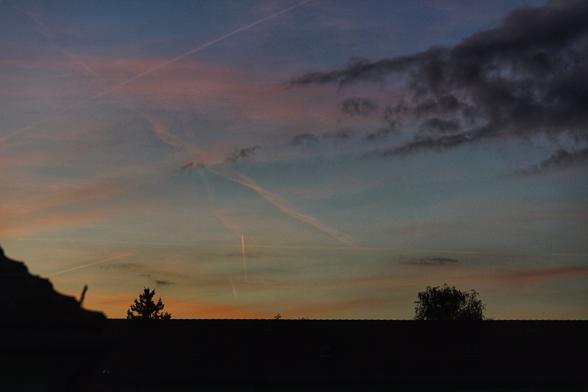 Orange bis rosa Wolken, daneben dunklere. Angeleuchteter Kondensstreifen, Dachspitze und Bäume
