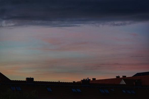 Wolkenbänder in unterschiedlichen Schattierungen, unten orange, dann rosa/rot, oben lila bis dunkelblau und schwarz.
Darunter Dächer