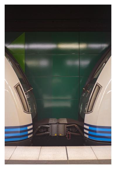 Foto im Hochformat. Die Kupplung zwischen zwei Waggons einer S-Bahn. Von beiden Waggons ist nur das Ende im Bild. Sie sind cremefarben, unten sind dunkel und hellblau Streifen. Davor sind die ebenfalls cremefarbenen Fliesen des Bahnsteigs. Hinter den Zügen ist eine Wand mit quadratischen grünen Kacheln. Darüber ist es schwarz. Das ganze Bild ist sehr symmetrisch aufgebaut und es spiegelt sich quasi in der Mitte.