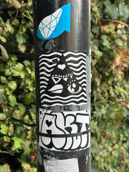 a sticker with a black and white abstract design, in the form of a black triangle filled with shapes in front of wavy horizontal lines, on a pole. Below it is a sticker spelling either AKT or ART in tag throwup lettering