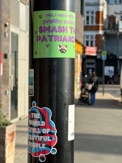 colourful stickers on a pole reading “yes I am pretty, pretty sure to smash the patriarchy” and “the world is full of beautiful people”