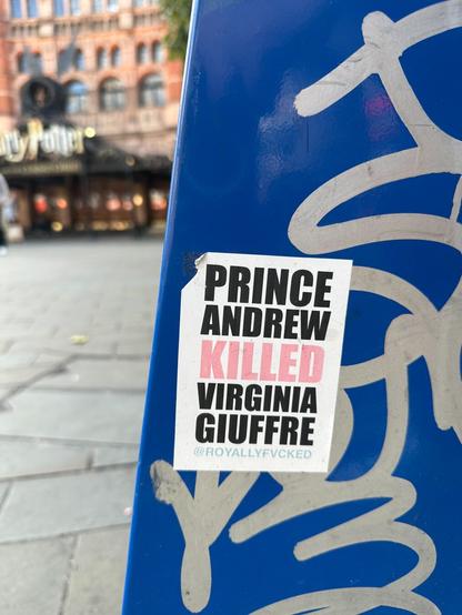 a sticker reading “PRINCE ANDREW KILLED VIRGINIA GIUFFRE” on a blue-painted surface covered with tags. In the background is what looks like a pub or eatery