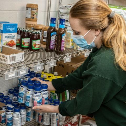 In a well-stocked store shelf, a person wearing a green shirt and a blue face mask is selecting items. The individual is holding a blue container and appears to be in the process of making a choice. The store is filled with various bottles and jars, each with different shapes, sizes, and colors, creating a vibrant display of choices.