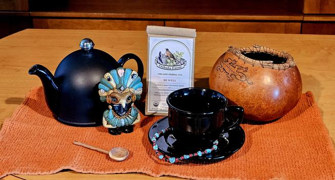 Tea tableau in desert colors commemorating Indigenous Peoples' Day. Upon a burnt orange cloth are a shiny black cup and saucer with a strand of turquoise, carnelian, black onyx,and silver beads entwined around it. Beside it is a carved obsidian statuette of an Aztec or Mayan eagle warrior encrusted with semiprecious stones. Behind them are a black teapot and a gourd basket etched with an iguana design. There is also a carved wooden spoon, and the tea is an herbal blend called Be Well from Oshala Farm in Oregon. It has elderberry, nettle, rosehip, burdock, echinacea, elderflower, and red clover.