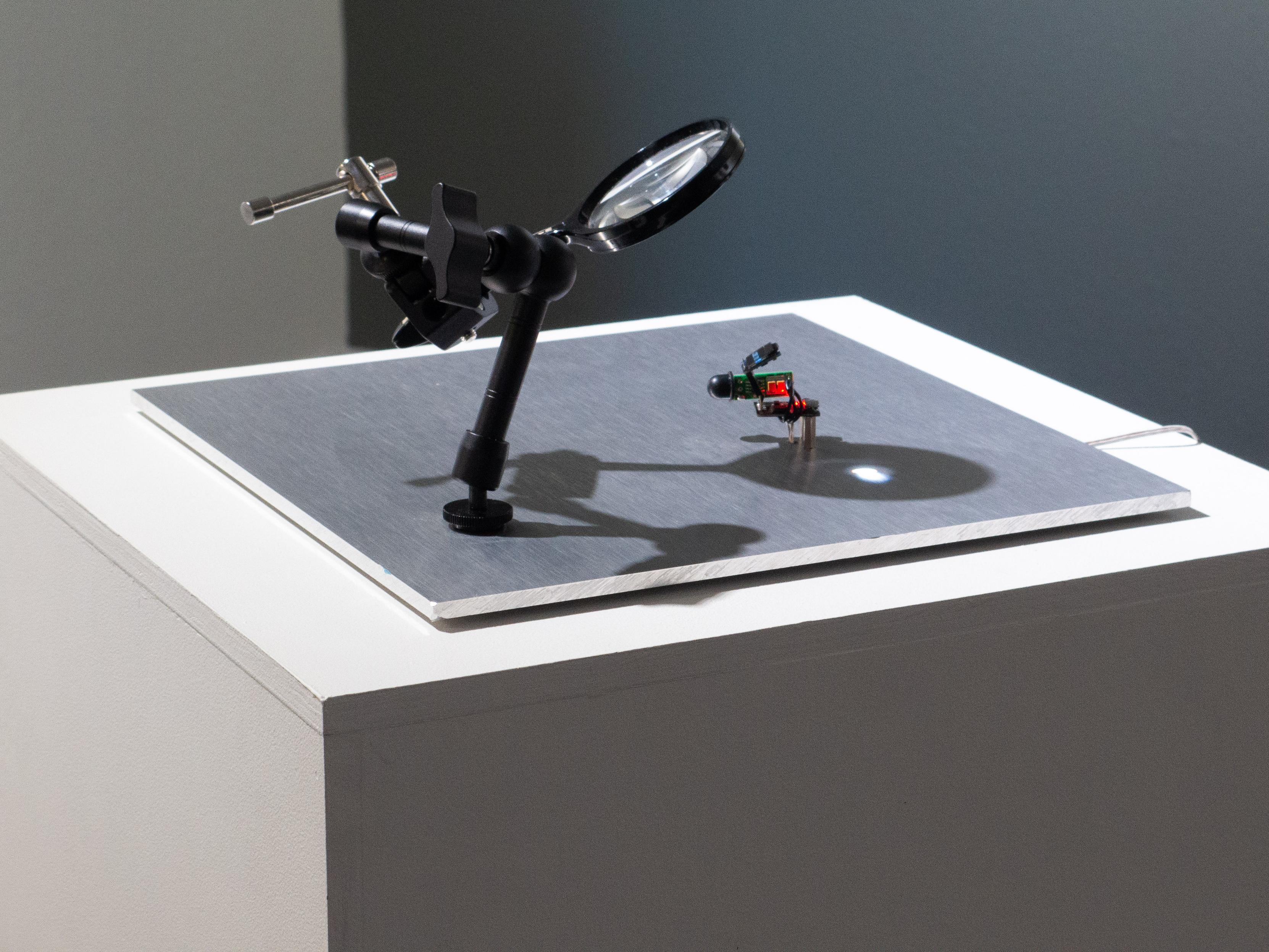 Perched on a square pedestal is a thin panel of brushed aluminum. It has an articulated arm mounted to it, which is holding a magnifying glass. The glass offers a peek at a tiny, precarious, glowing device.