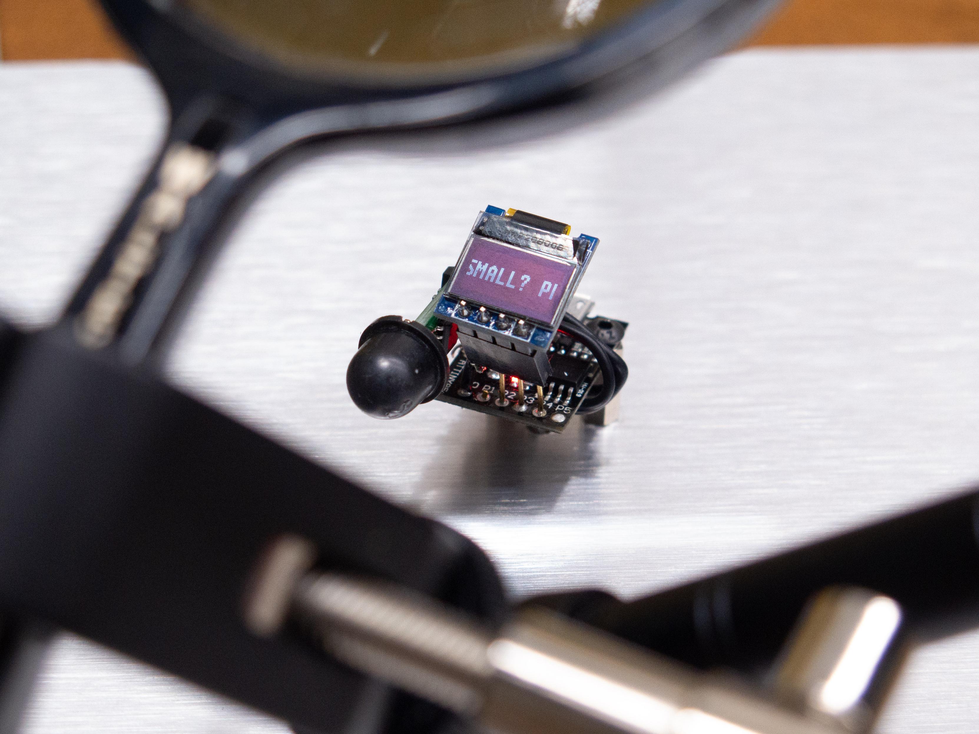 This closer view shows a little more of the device, which has a tiny screen (~0.49in / 12mm diagonal viewable area; 64x32 pixels) reading a partial message: "SMALL? P..." It is attached to a small microcontroller with a motion sensor. The magnifying glass looms around the edges of the frame.
